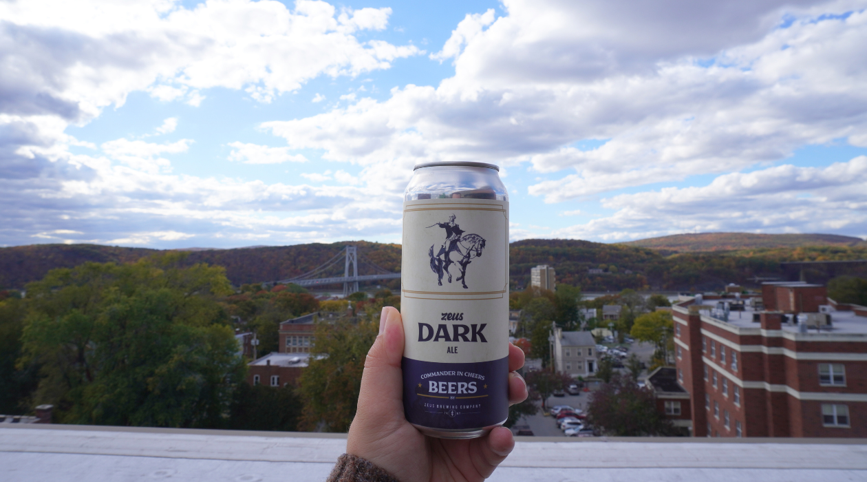 A can of Zeus Dark Ale is held up by a hand on the rooftop overlooking the silver mid hudson bridge the blue hudson river and the colorful fall foliage on the mountains across the river